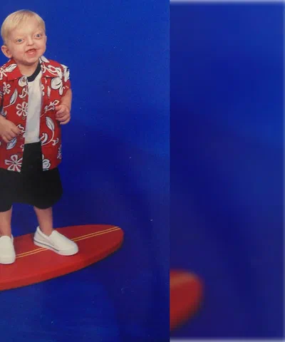 Alex as a toddler standing on a toy surfboard in a red Hawaiian shirt