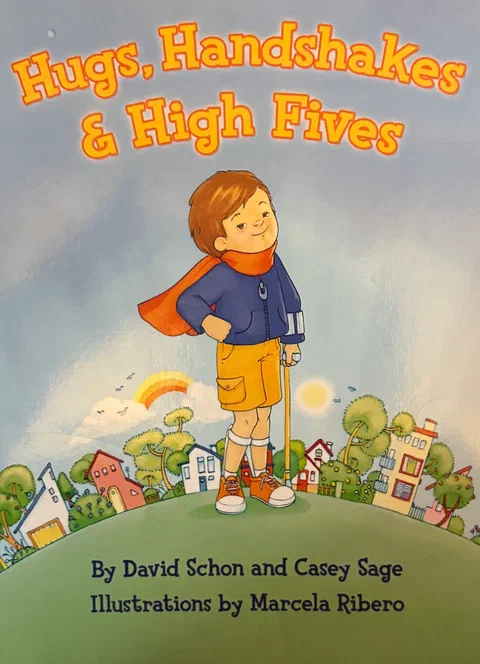 Cover of Hugs, Handshakes & High Fives: Travis, a boy in a red cape, gives a thumbs up on a hill above a town