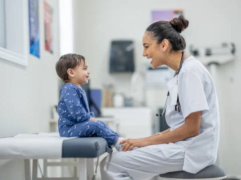 A toddler smiling at a pediatric check-up while a clinician listens with a stethoscope