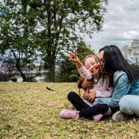 A mother embracing her laughing daughter, who has Down syndrome, while they sit together on the grass